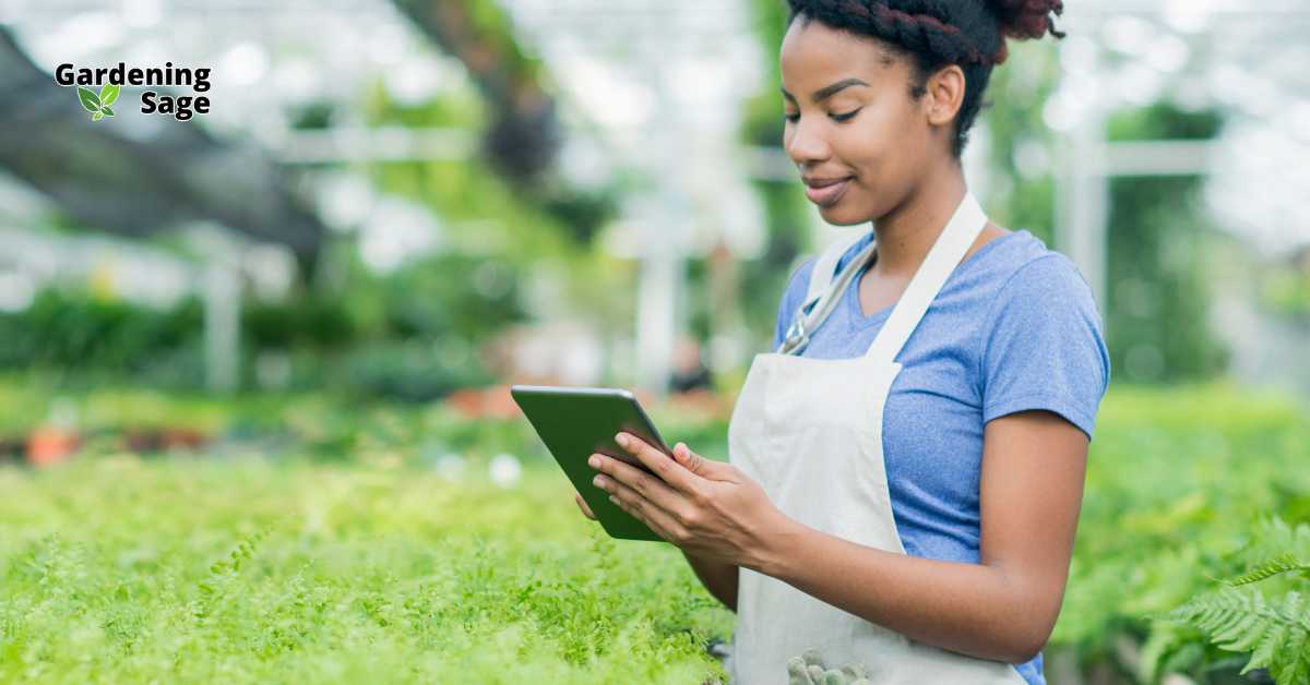 The image depicts a modern gardening scene where a young female gardener, dressed in a casual blue t-shirt and white apron, is using a tablet in a lush, well-maintained greenhouse. Her focused expression suggests she is likely researching or recording details about plant care, showcasing the integration of technology in gardening. This setting reflects a blend of traditional gardening with modern technological advancements, emphasizing how digital tools can enhance the efficiency and knowledge in managing a garden. The greenhouse environment around her is vibrant and full of various plants, indicating a healthy, thriving garden that benefits from precise and informed care practices.