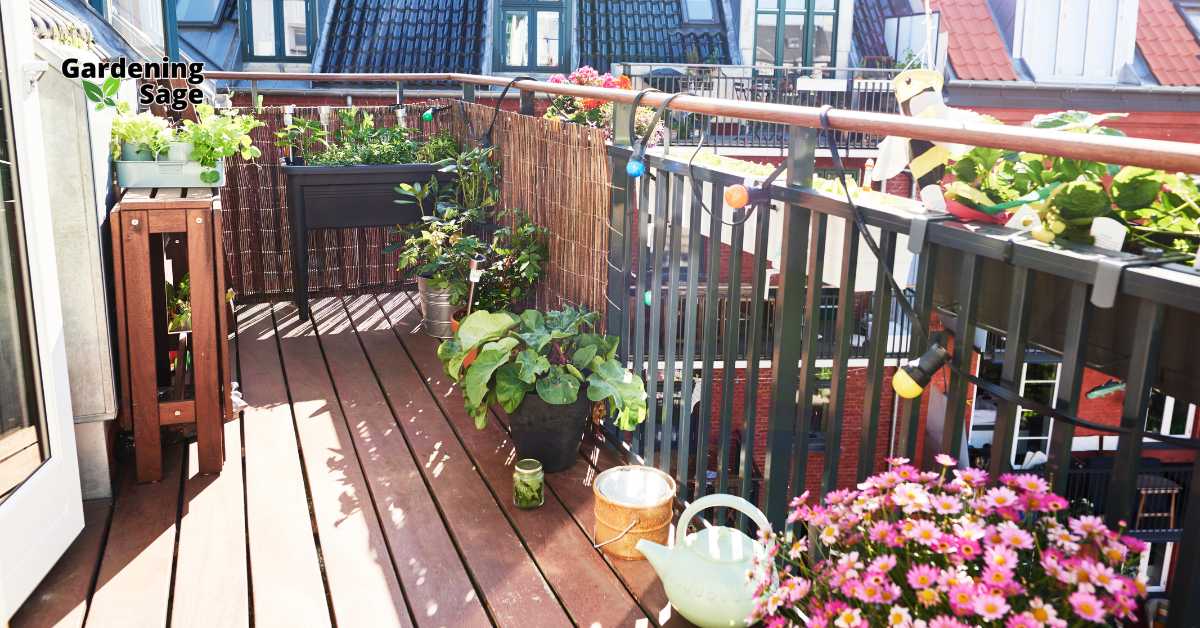 This image depicts a small, well-utilized balcony garden in an urban setting. The garden features a variety of potted plants, including flowers, herbs, and leafy greens, arranged neatly along the wooden decking. A small raised planter is positioned against a bamboo privacy screen that lines the balcony railing. The space is adorned with colorful string lights, adding a cozy atmosphere to the garden. A watering can and a few gardening tools are also visible, indicating an active and well-maintained urban garden. The overall setup exemplifies efficient use of limited space for gardening in a compact area.